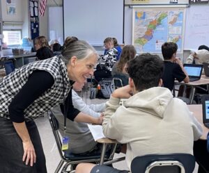 A teacher helps a student work at their desk with a smile