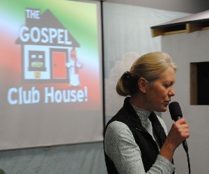 An adult speaks into a microphone as she leads a children's ministry service.